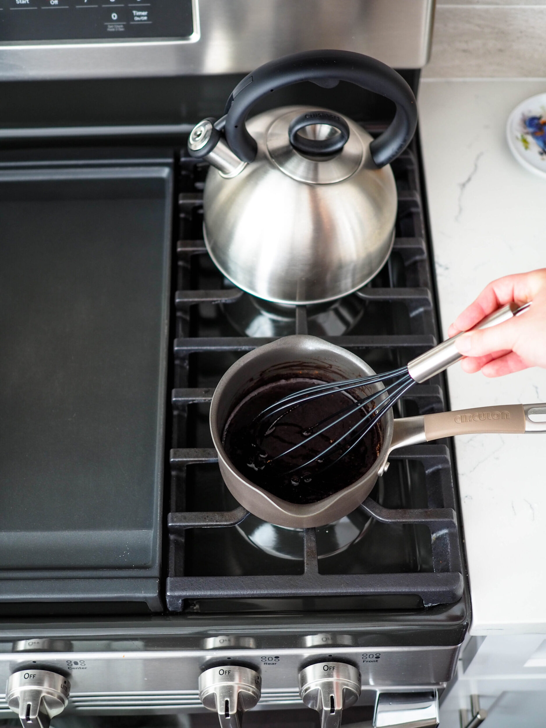 A whisk in a small saucepan filled with dark brown liquid on the stove.