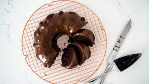 A chocolate Bundt cake on a copper wire rack with a few pieces cut up and laid in the ring of the cake.