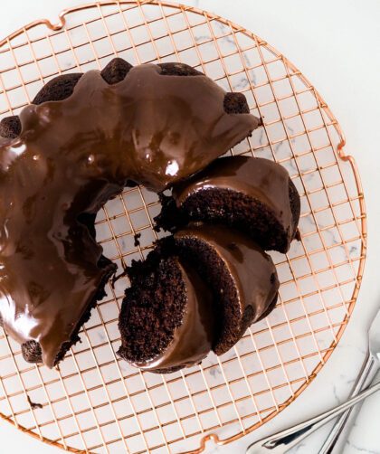 A chocolate Bundt cake on a copper wire rack with a few pieces cut up and laid in the ring of the cake.