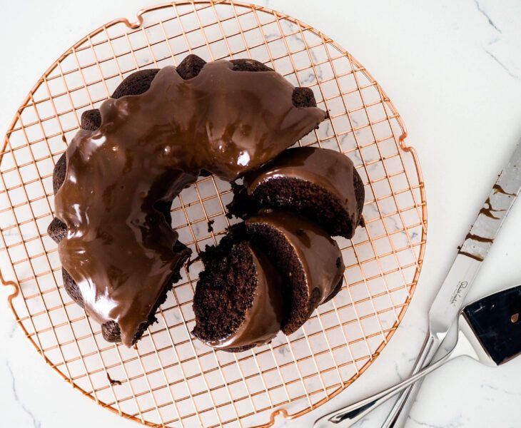 A chocolate Bundt cake on a copper wire rack with a few pieces cut up and laid in the ring of the cake.