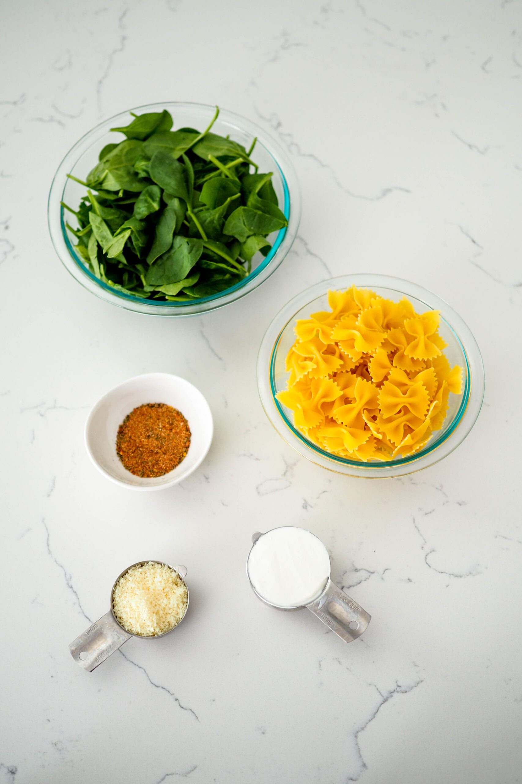 Spinach, pasta, sour cream, Parmesan cheese, and Cajun seasoning on a white quartz countertop.