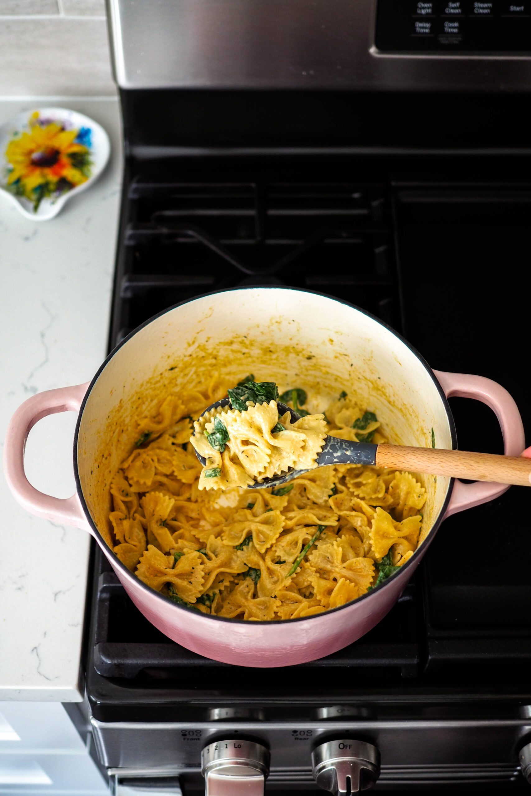 A wooden spoon holds up a bite of creamy Cajun spinach pasta out of a Dutch oven on the stove.