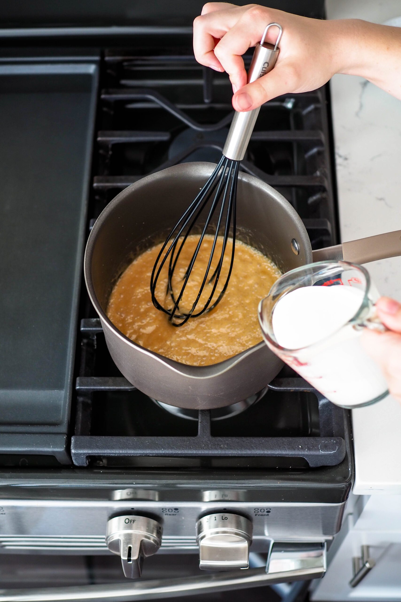 A measuring cup with a pour spout is poised over the edge of a pot with caramel.