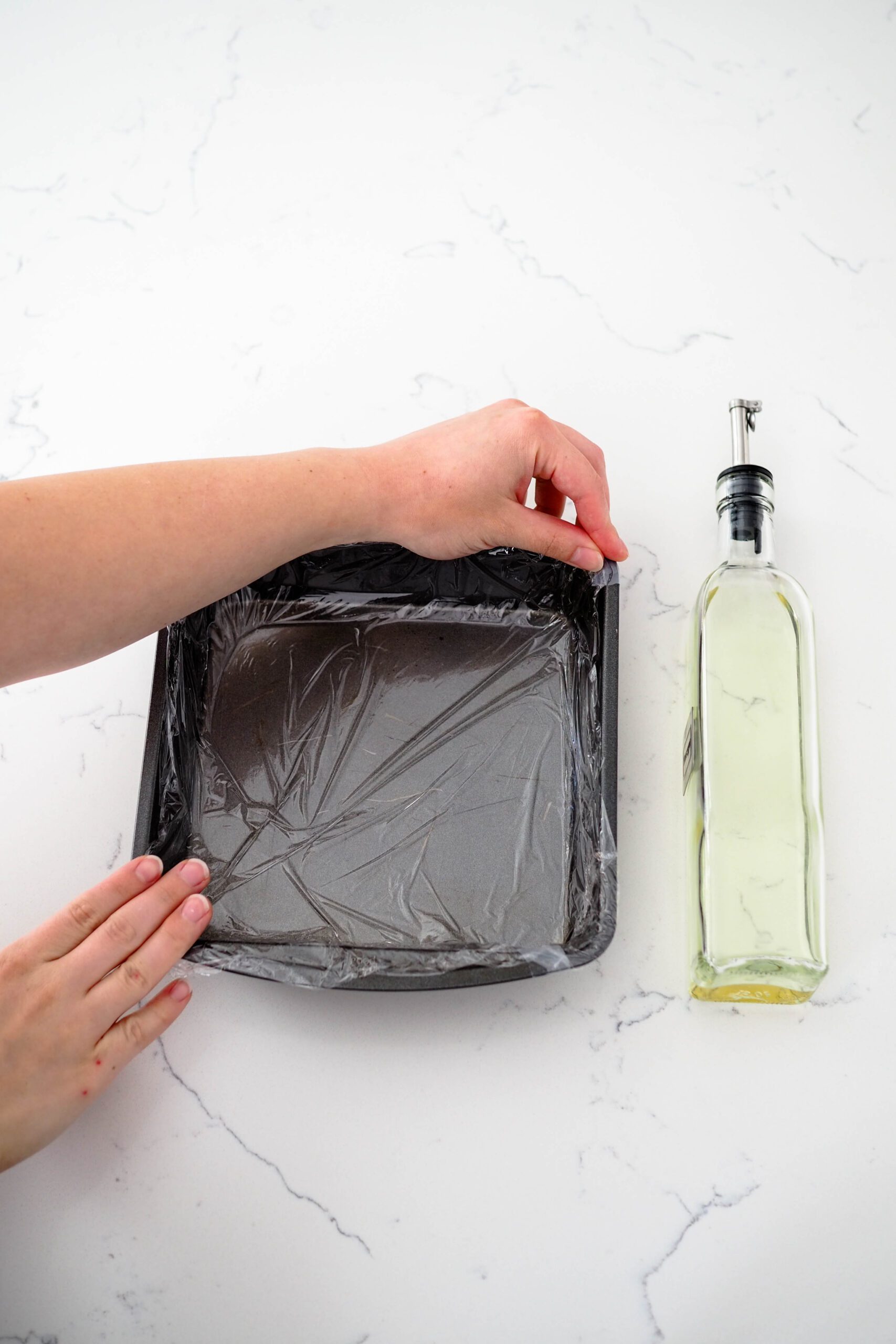 Two hands prepare a baking pan lined with plastic wrap.