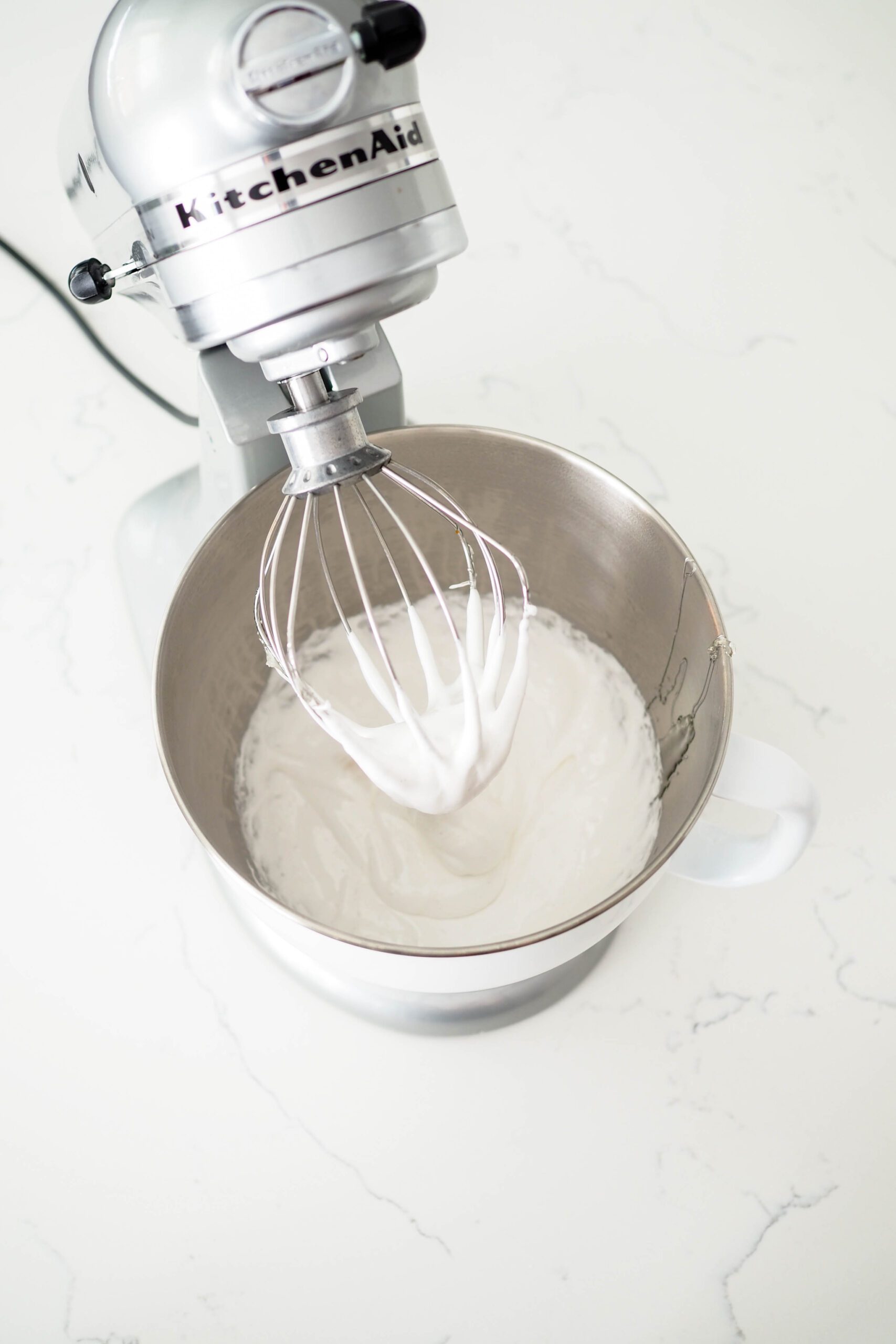 Marshmallow gathers in the whisk of a stand mixer.