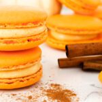 Pumpkin spice macarons are stacked next to cinnamon sticks.