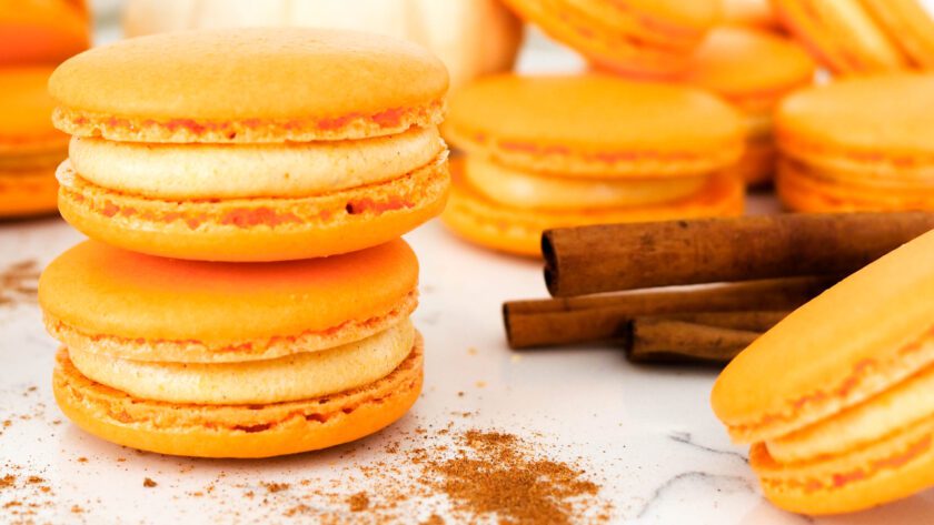 Pumpkin spice macarons are stacked next to cinnamon sticks.
