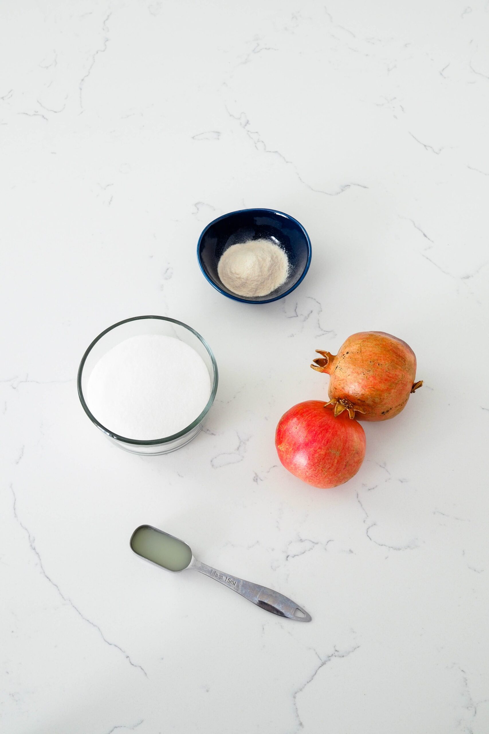 Two pomegranates, lemon juice, sugar, and pectin on a white counter.