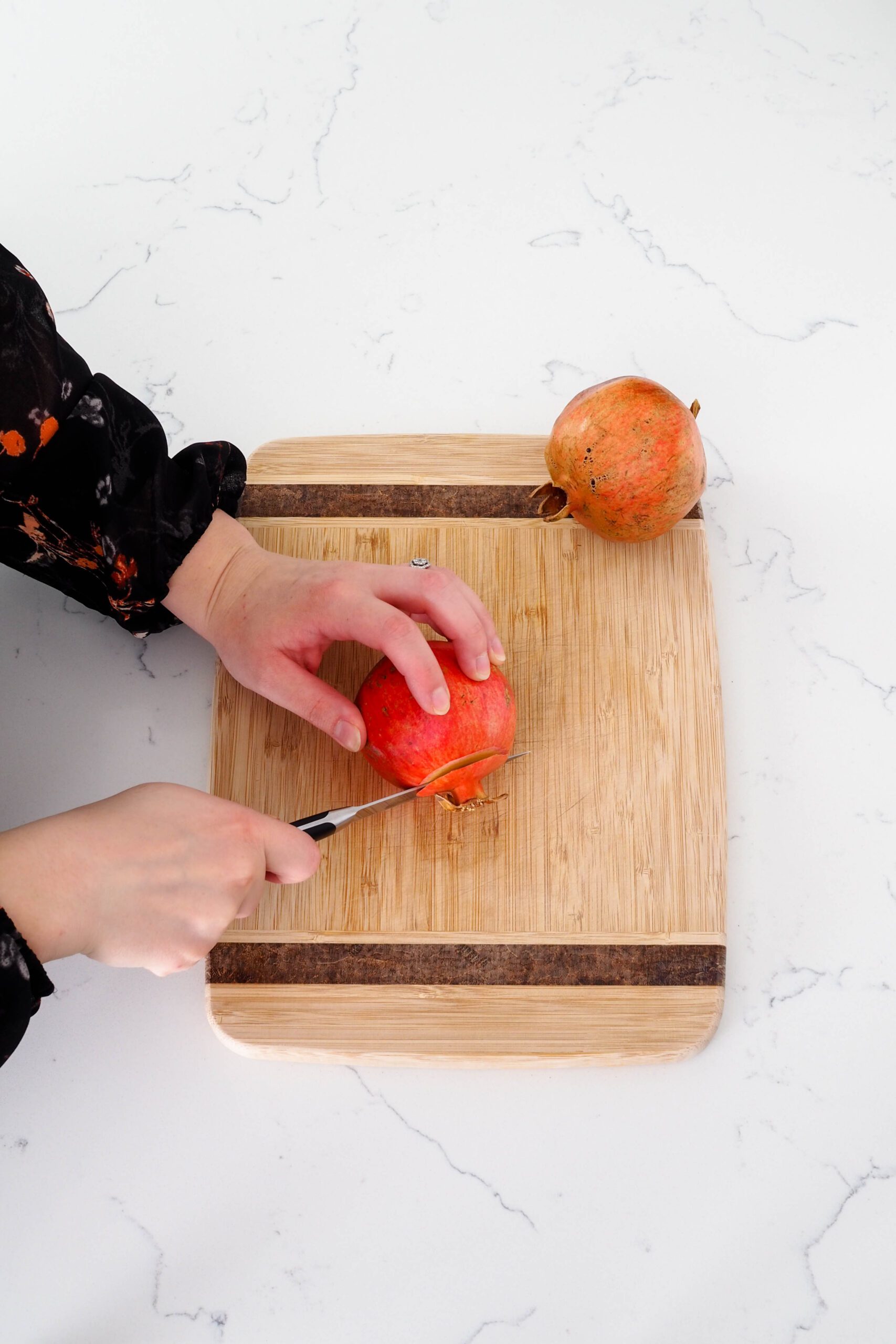 The crown of a pomegranate is being sliced off on a cutting board with a paring knife.