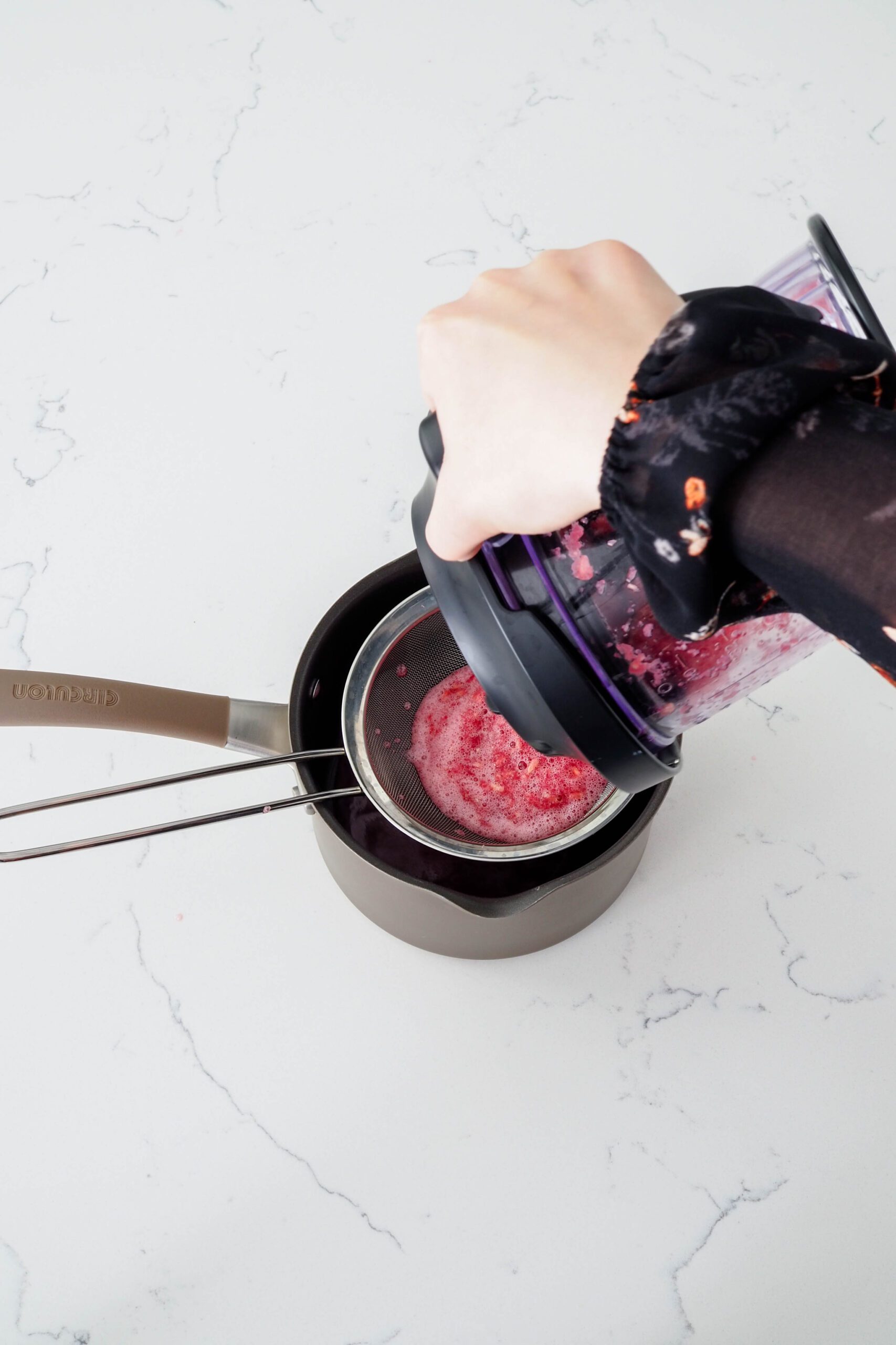 A hand pours pomegranate juice into a fine mesh strainer over a pot.