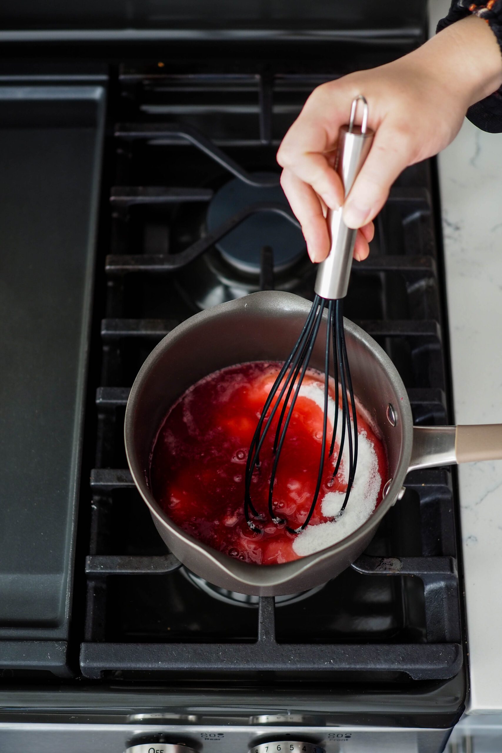 A whisk stirs sugar into pomegranate jam over the stove.