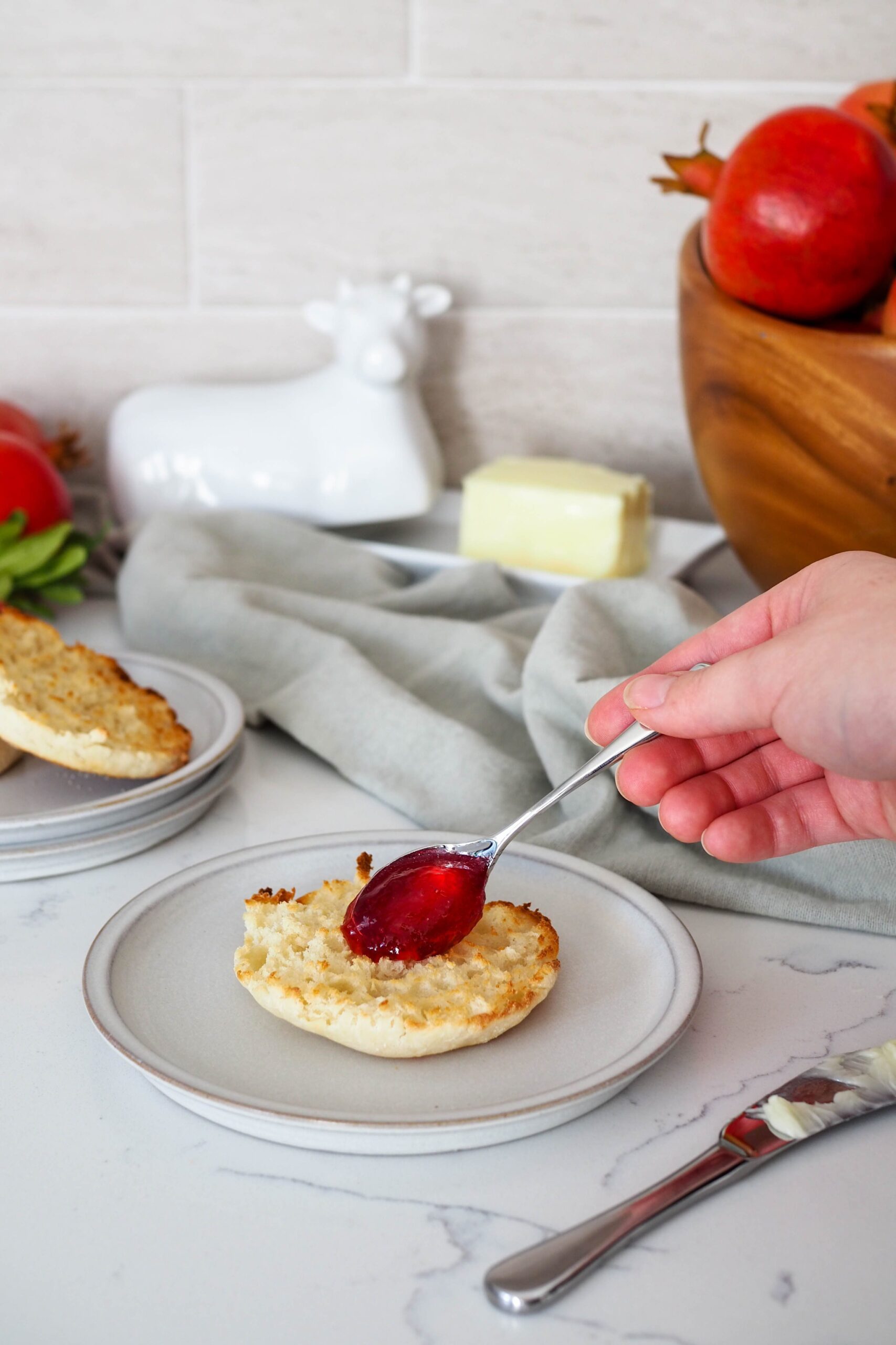 A small spoon spreads pomegranate jelly onto an English muffin.