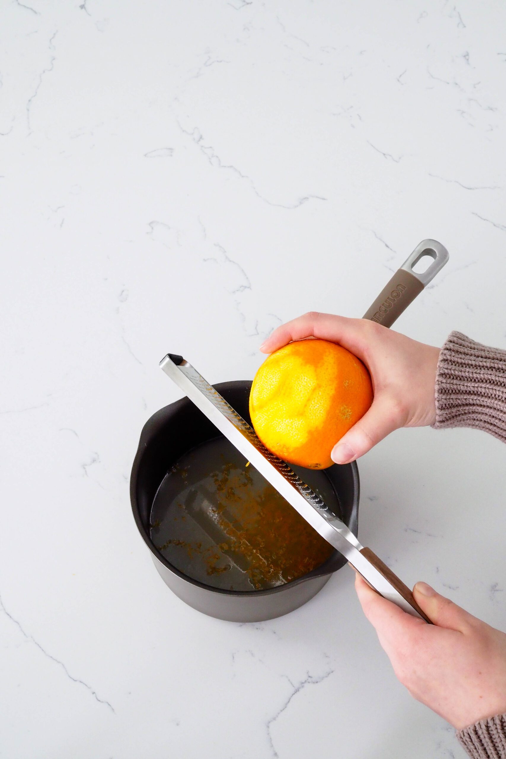 Two hands zest an orange directly over a pot filled with water and sugar.
