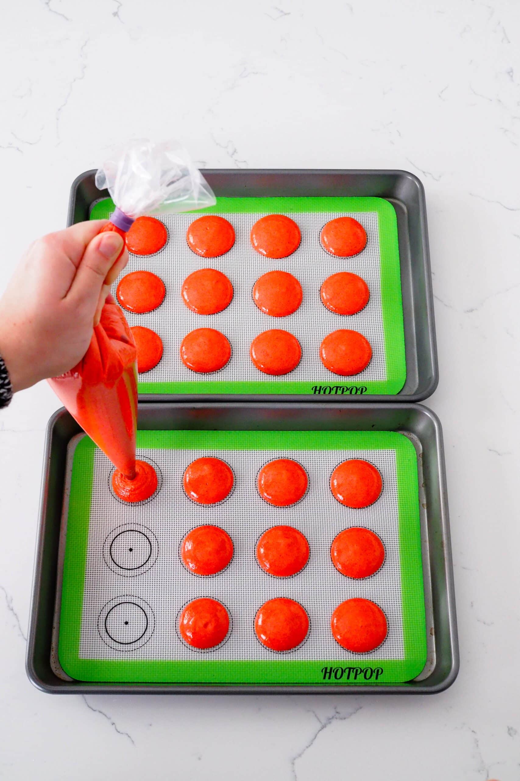 A hand pipes red macaron batter into circles on a silicone macaron mat.
