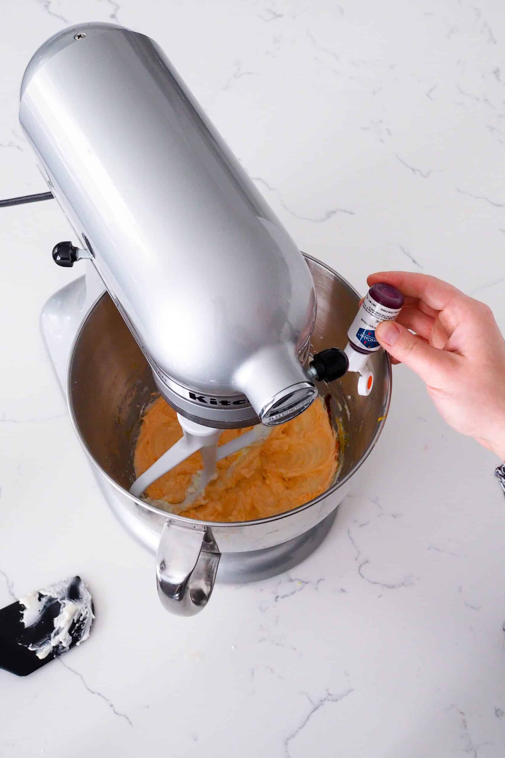 A hand squeezes a drop of orange food coloring into a mixing bowl with orange buttercream.