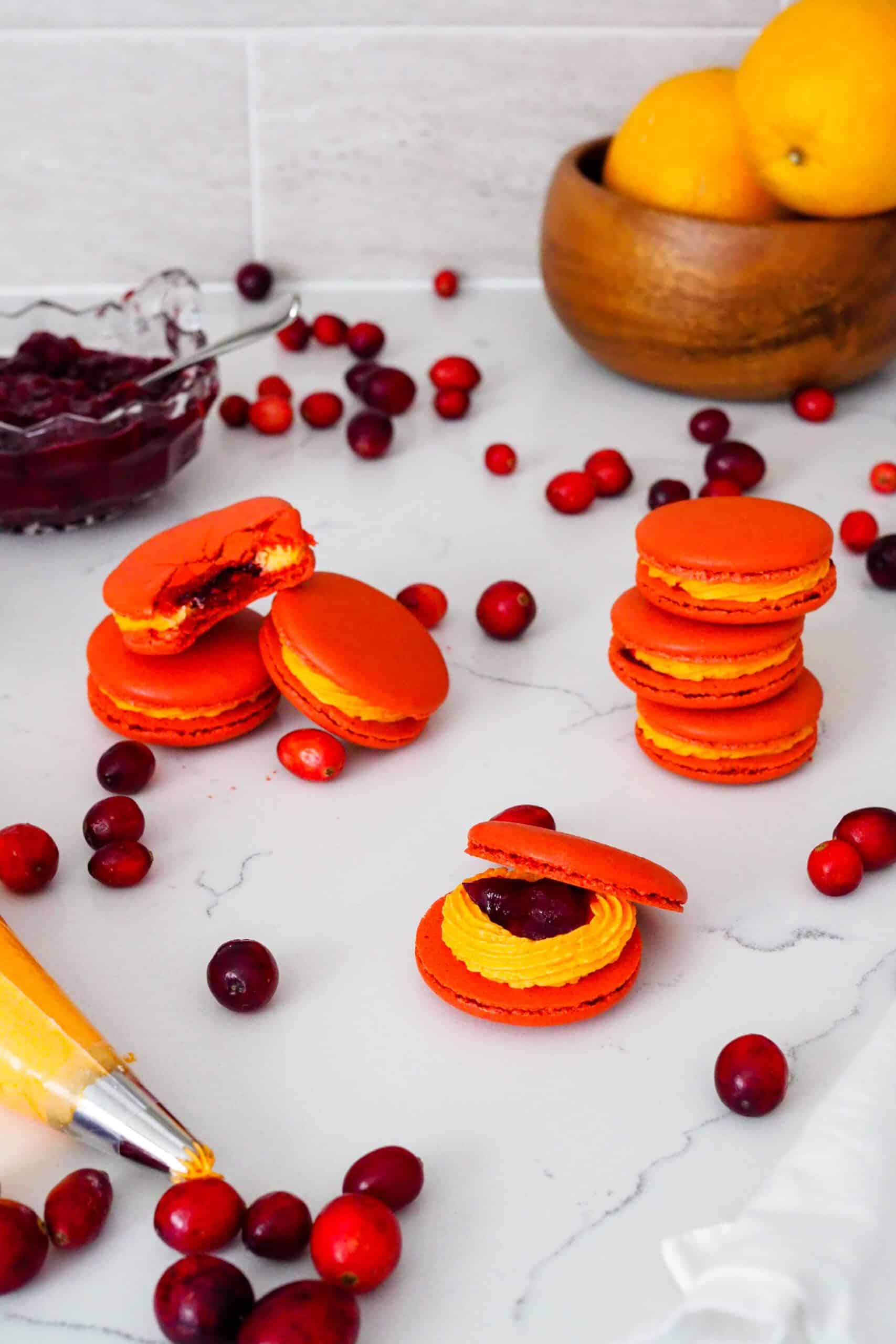 Red and orange macarons are stacked on a white counter with cranberries surrounding them.