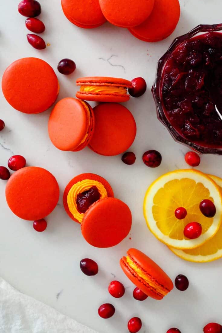A stack of red and orange macarons near orange slices and a bowl of cranberry sauce.