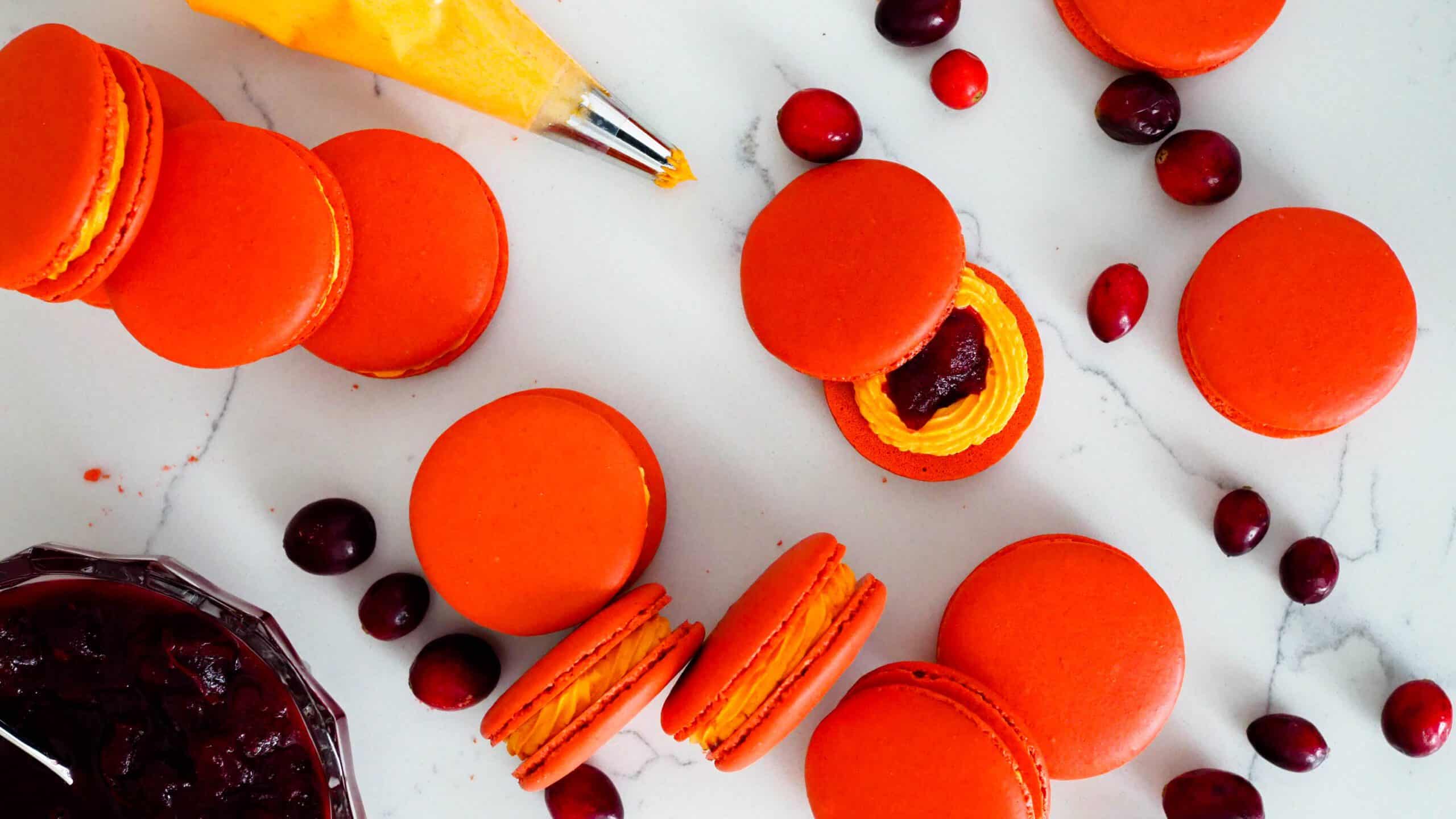 Cranberry orange macarons are arranged artfully on a counter with cranberries, a piping bag, and a bowl of cranberry sauce.