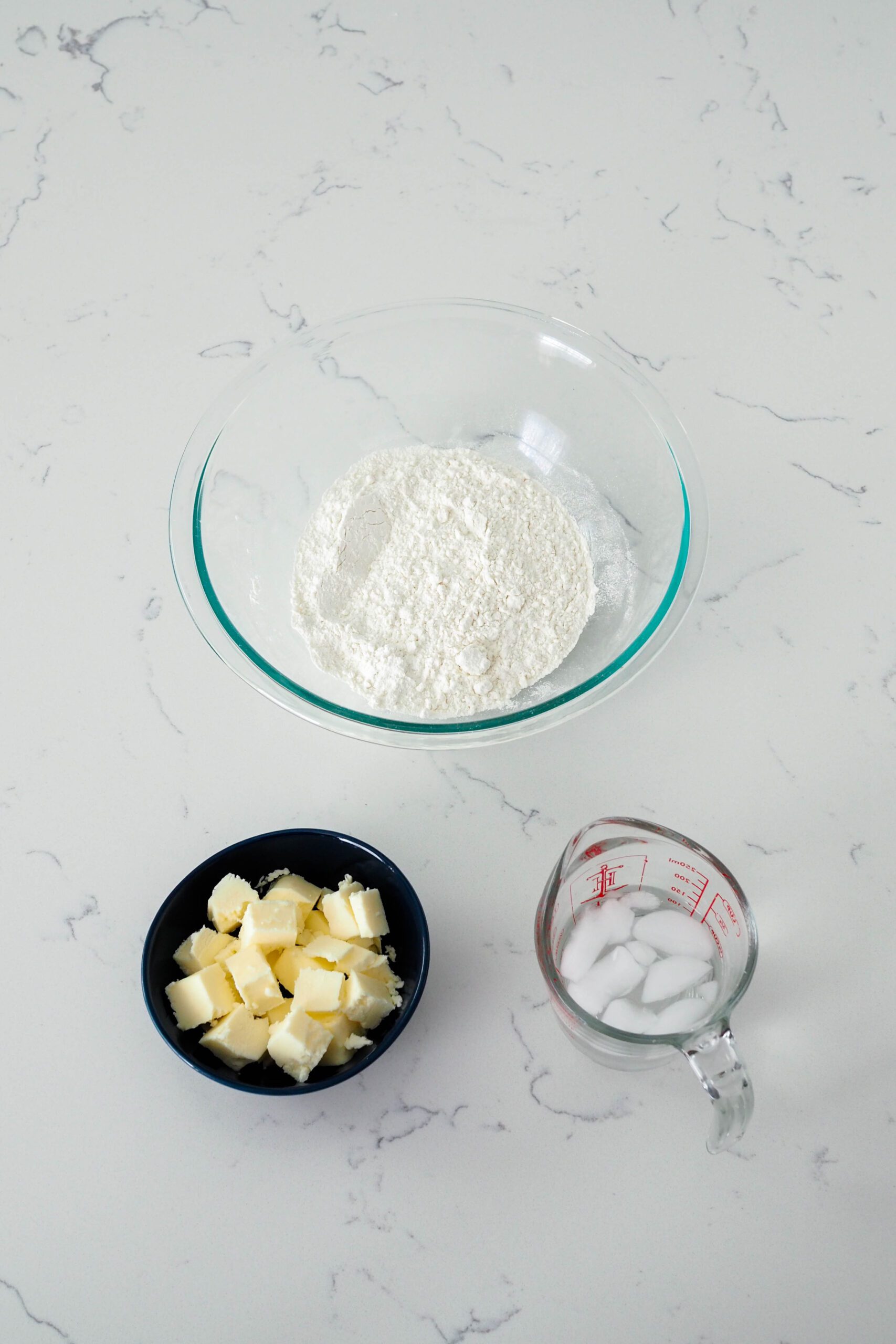 A big bowl of flour, a small bowl of butter, and a measuring cup with water are on a quartz counter.