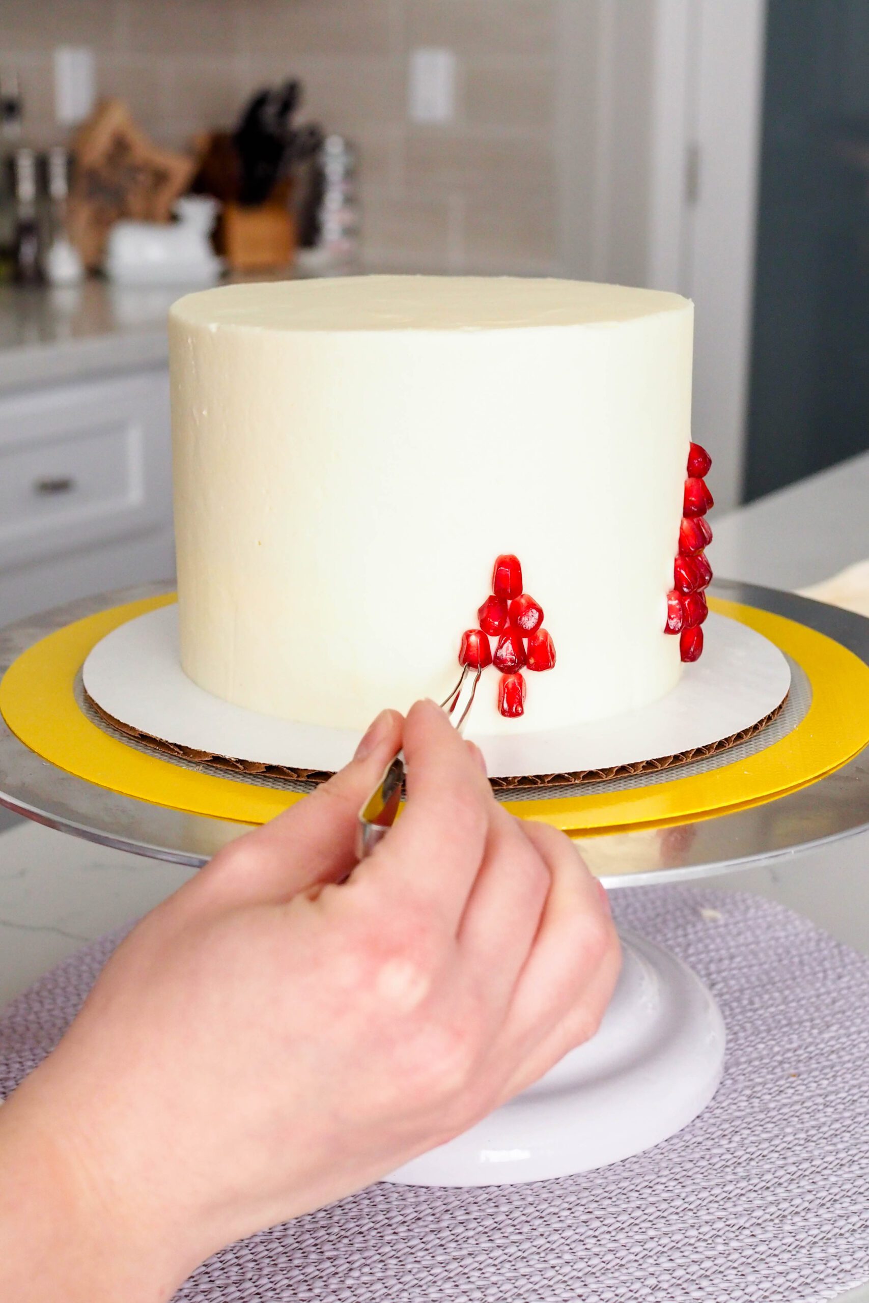 A hand uses decorating tweezers to place pomegranate arils on the side of a cake.
