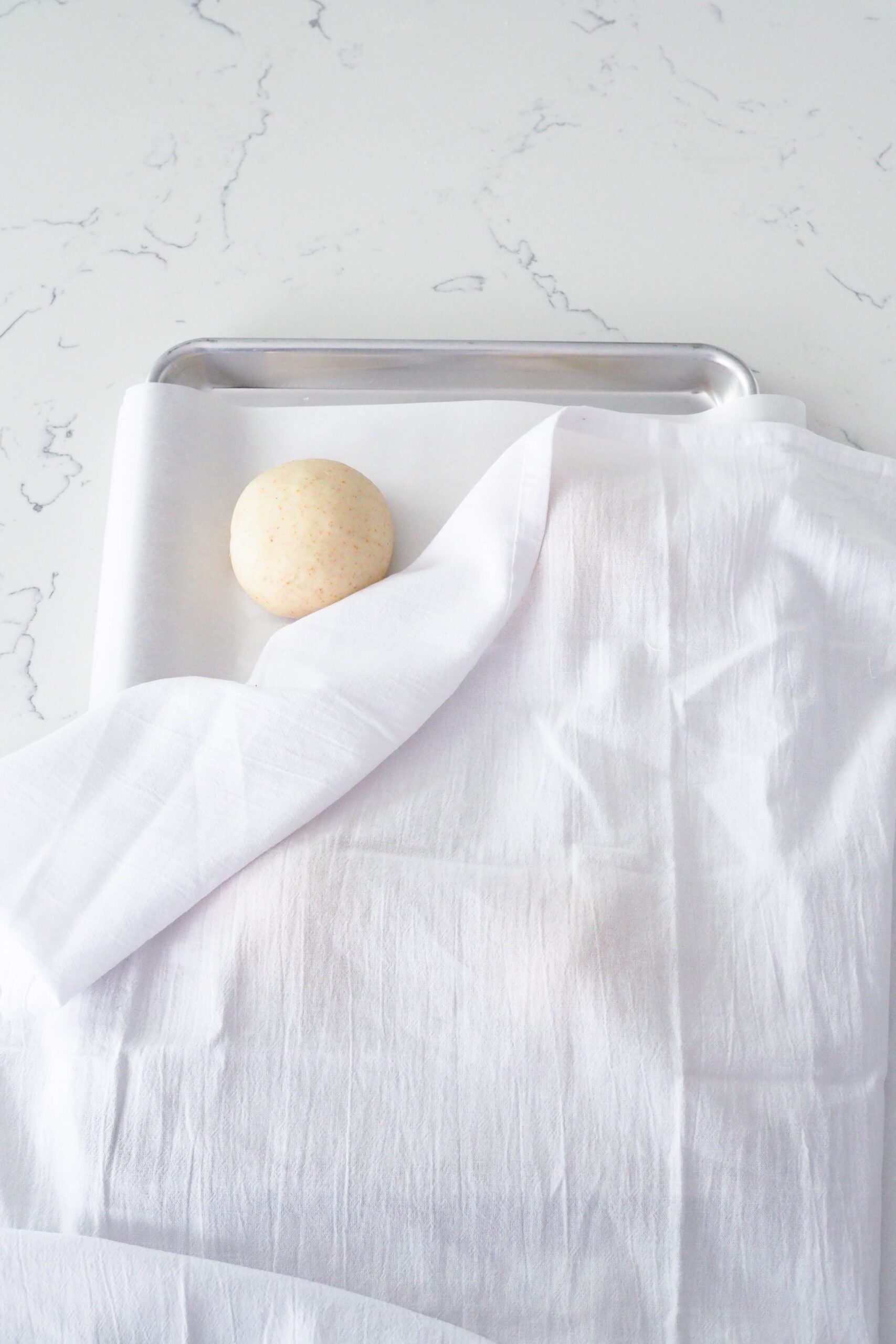 A dough ball peeks out behind a white kitchen towel on a baking sheet.