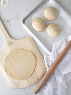 A rolled out pizza crust on a pizza peel, with three small dough balls and a rolling pin beside it.