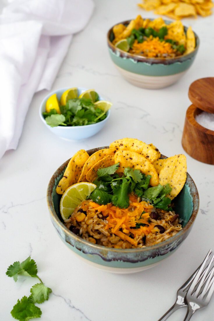 A bowl of one-pot chicken and brown rice with black beans and corn topped with tortilla chips, cilantro, lime, and cheese.
