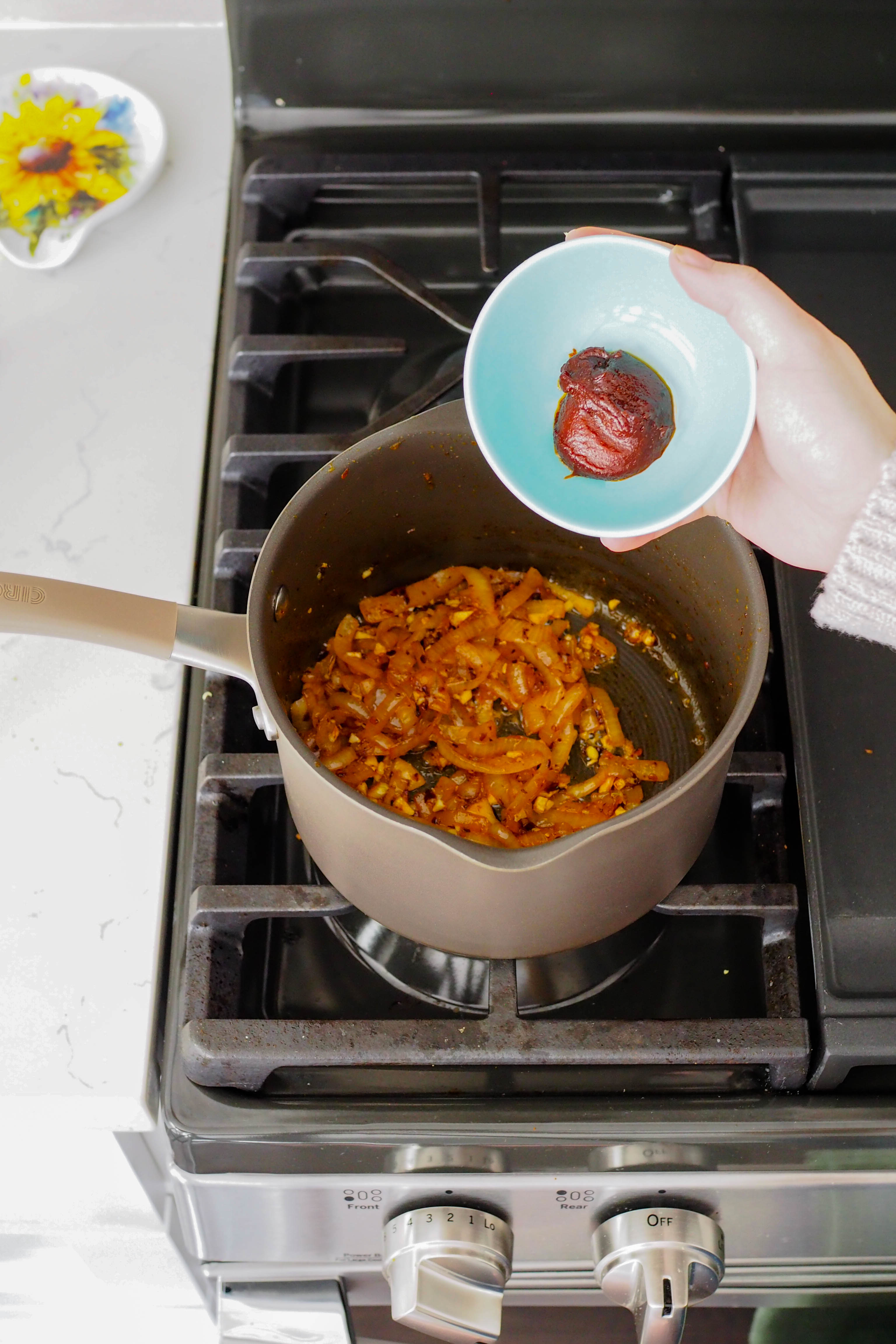 A hand pours gochujang into a pot of caramelized onions.