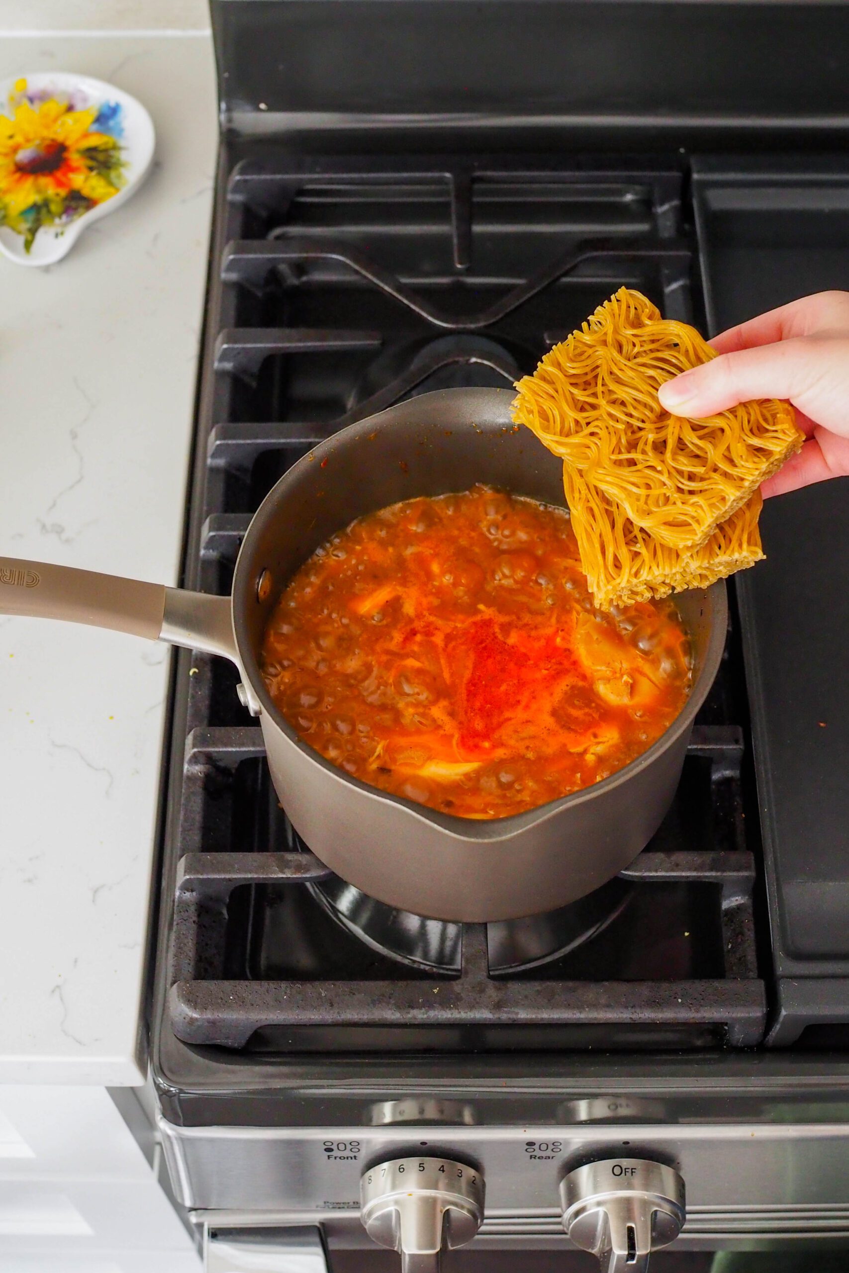 A hand places two cakes of dried ramen noodles into ramen broth.