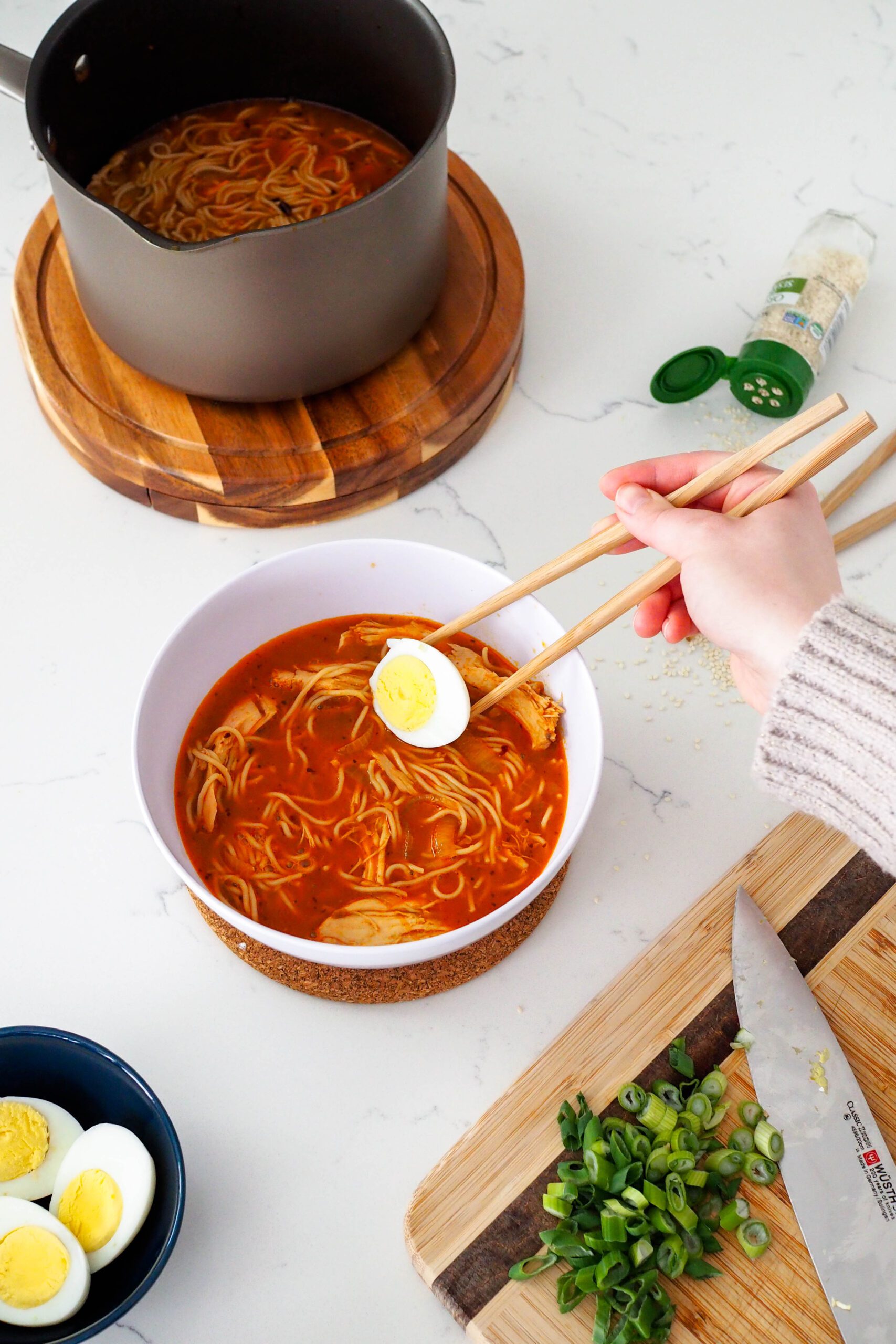 A hand places half of a boiled egg into a bowl of ramen with chopsticks.