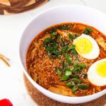 A bowl of gochujang chicken ramen with two halves of a boiled egg in it.