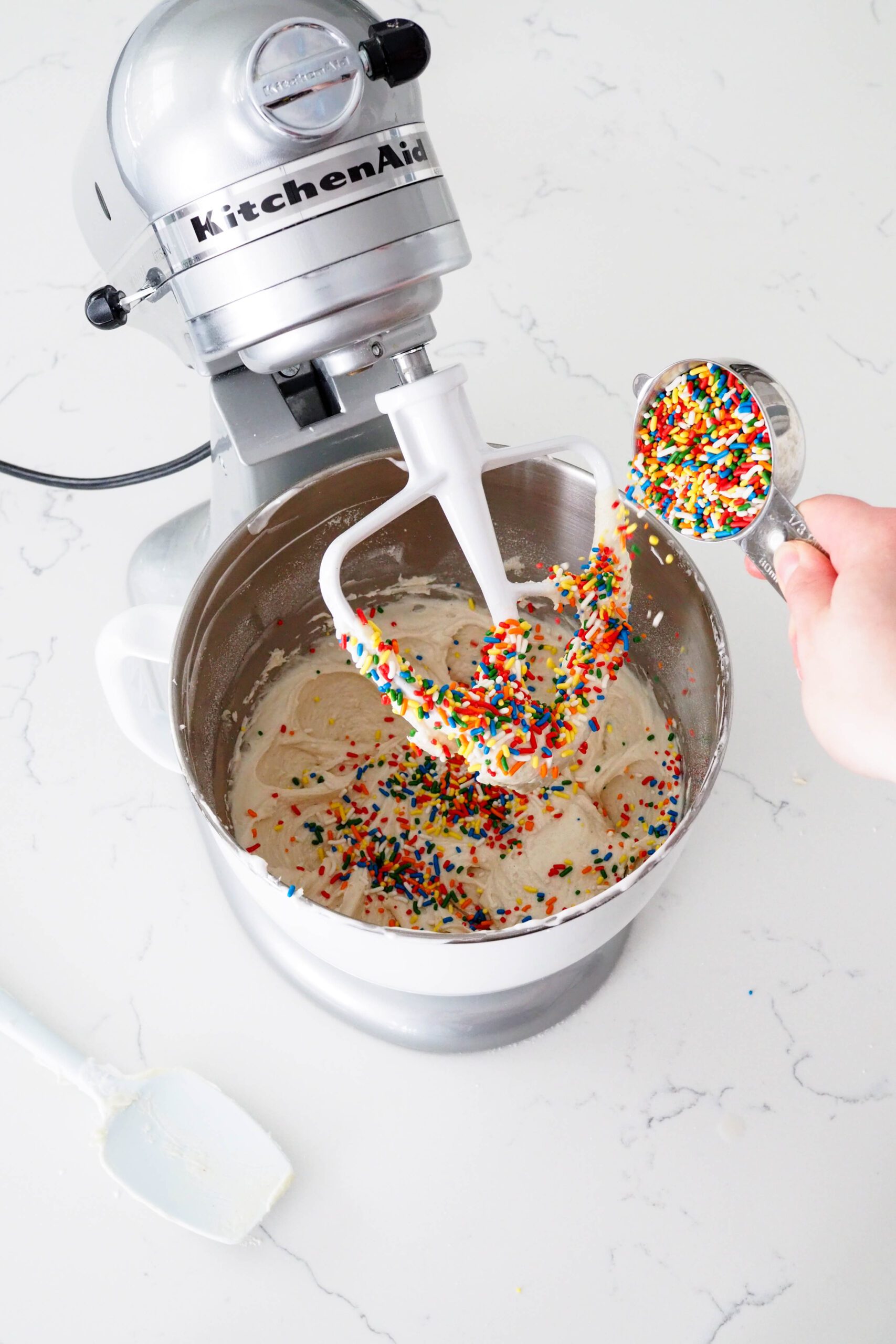 A measuring cup pours sprinkles into a thick mini confetti layer cake batter.