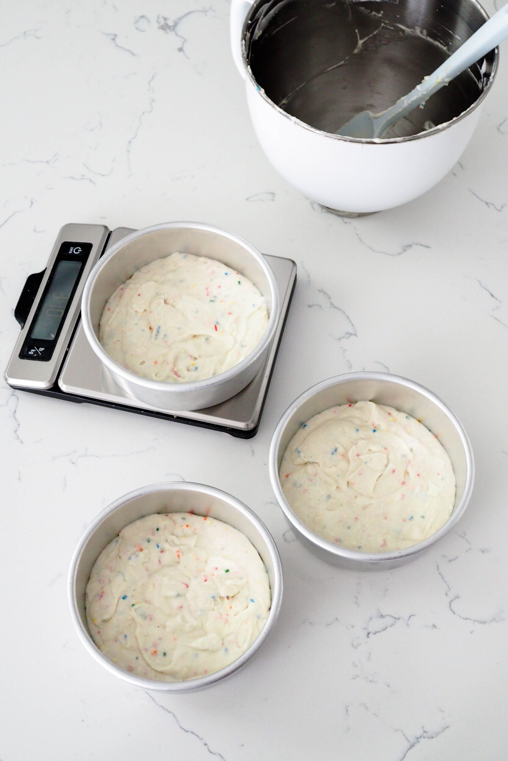 Three cake pans are filled with sprinkle cake batter. One rests on a digital scale.