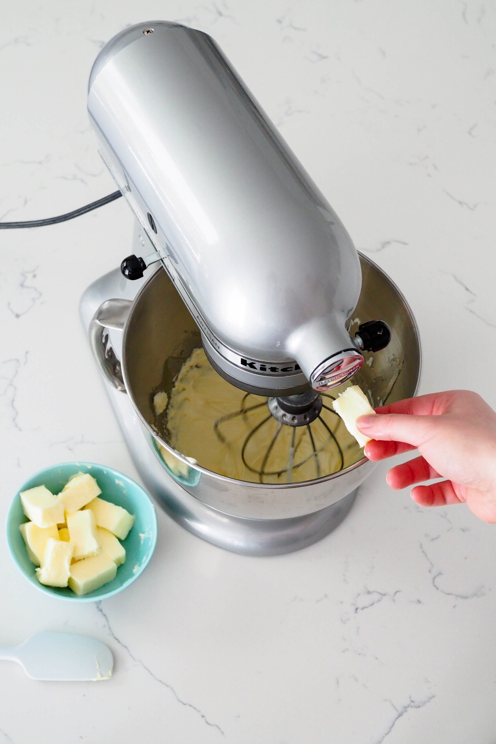 A hand holds a small piece of butter in front of a mixer with a whisk attachment.