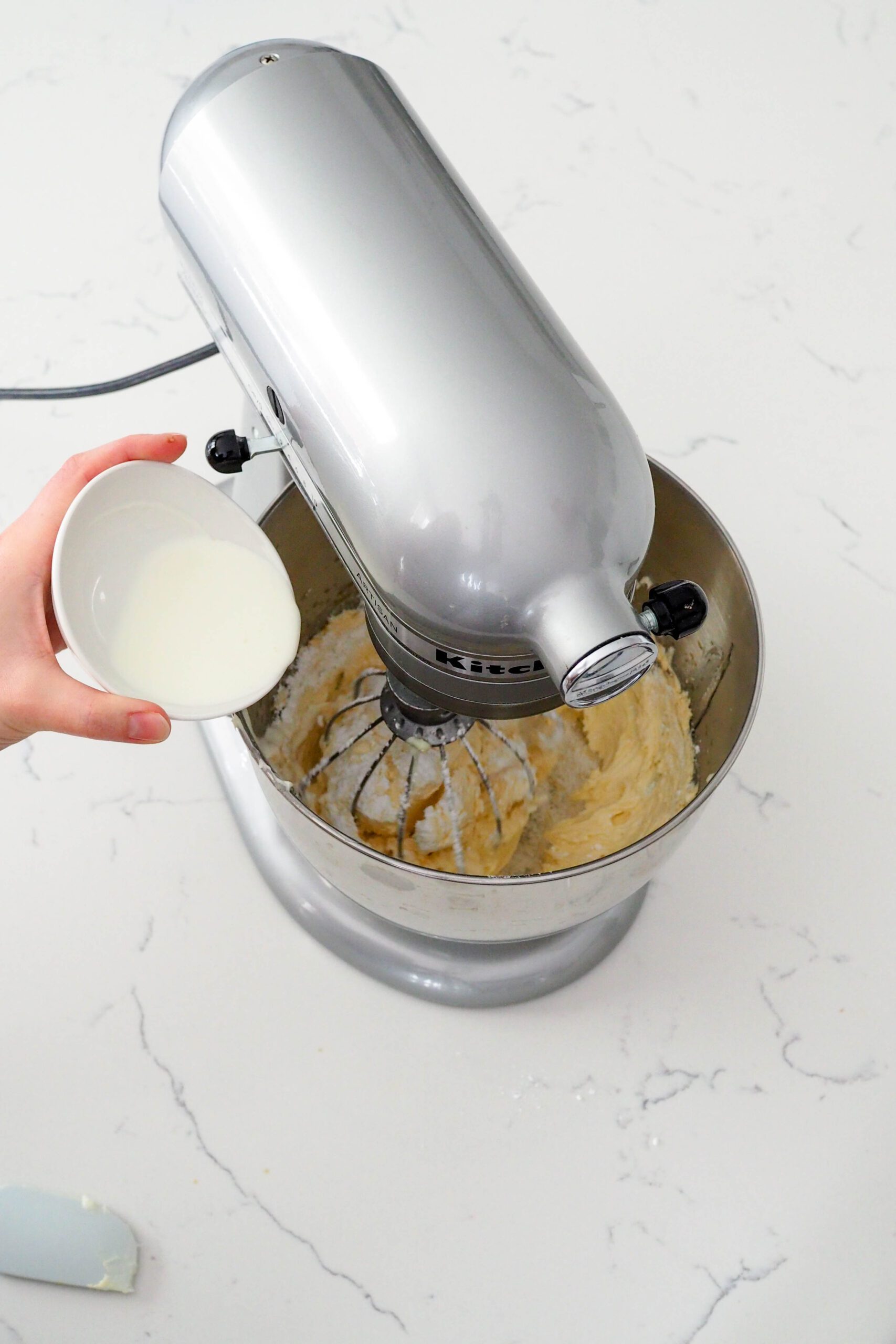 A hand pours heavy cream into French buttercream in a mixer.