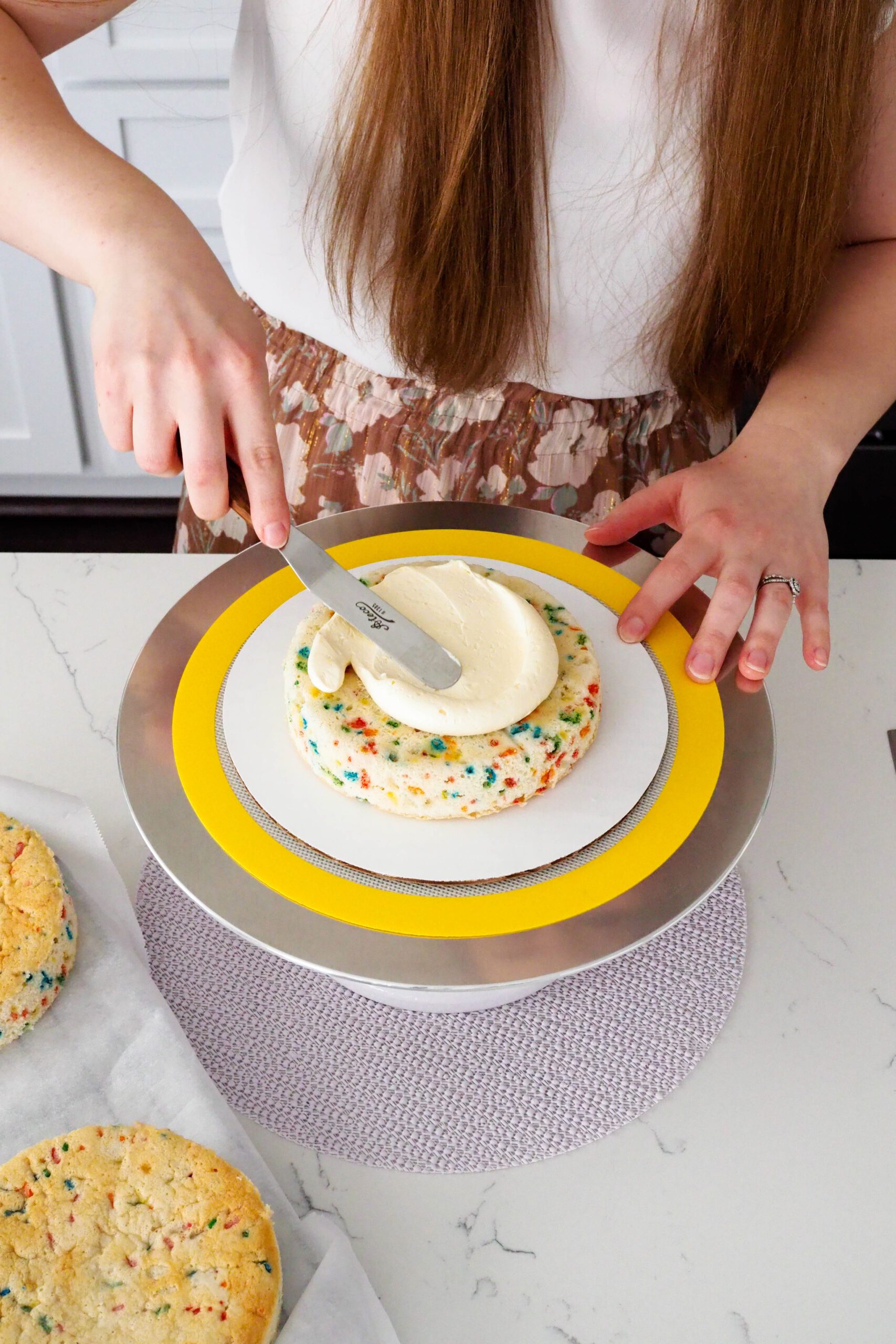 A woman spreads French buttercream over a mini confetti layer cake on a turntable.