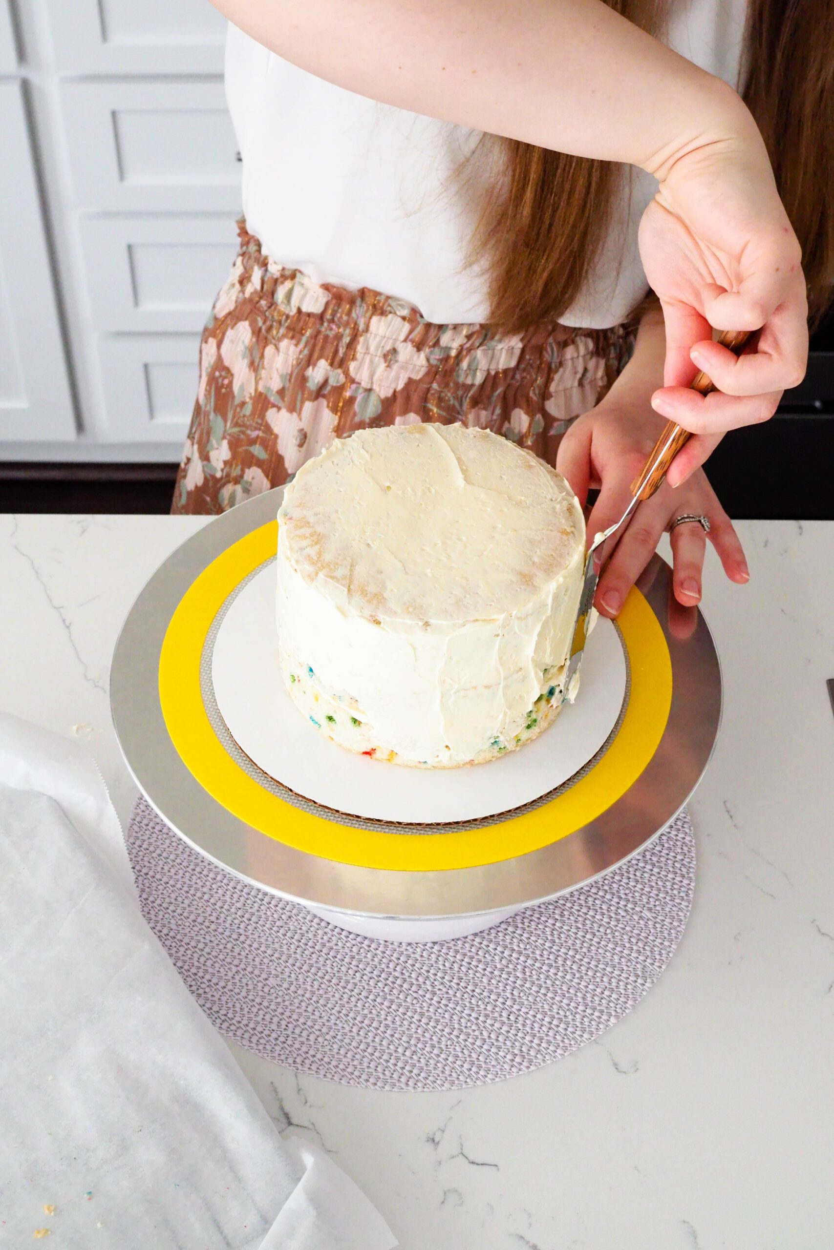 A woman smooths buttercream over the sides of a mini confetti layer cake.