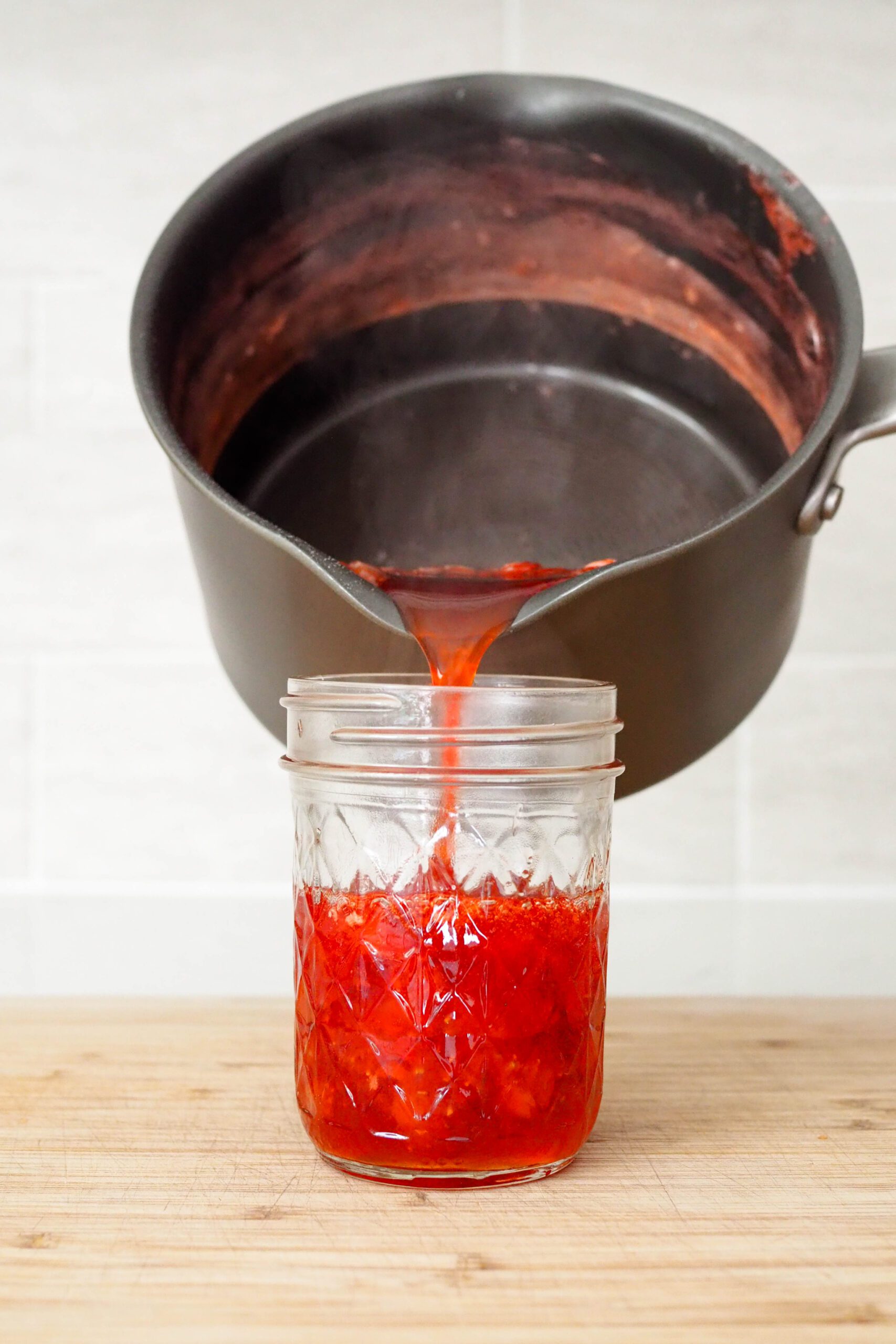 A saucepan pours strawberry champagne jam into a jar.