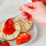 A hand spreads strawberry champagne jam on an English muffin.