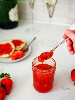 A hand scoops strawberry champagne jam out of a jar. Two champagne glasses stand behind it.