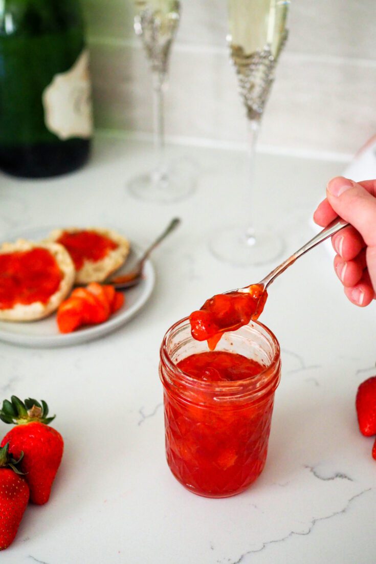 A hand scoops strawberry champagne jam out of a jar. Two champagne glasses stand behind it.