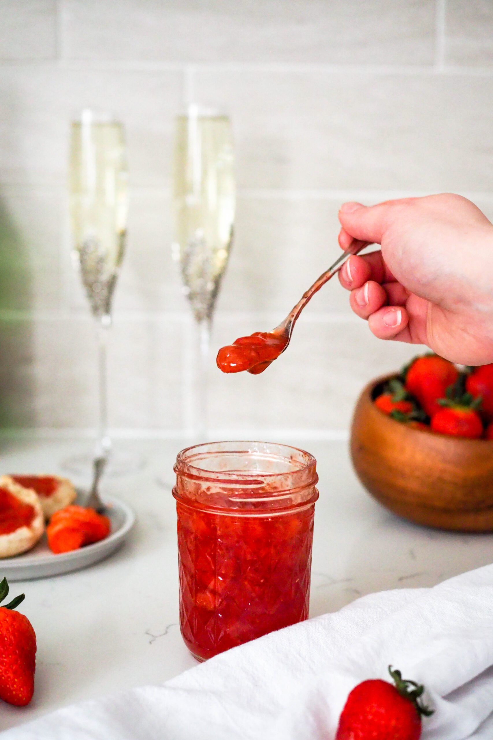 A hand scoops strawberry champagne jam out of a jar. Two champagne glasses stand behind it.