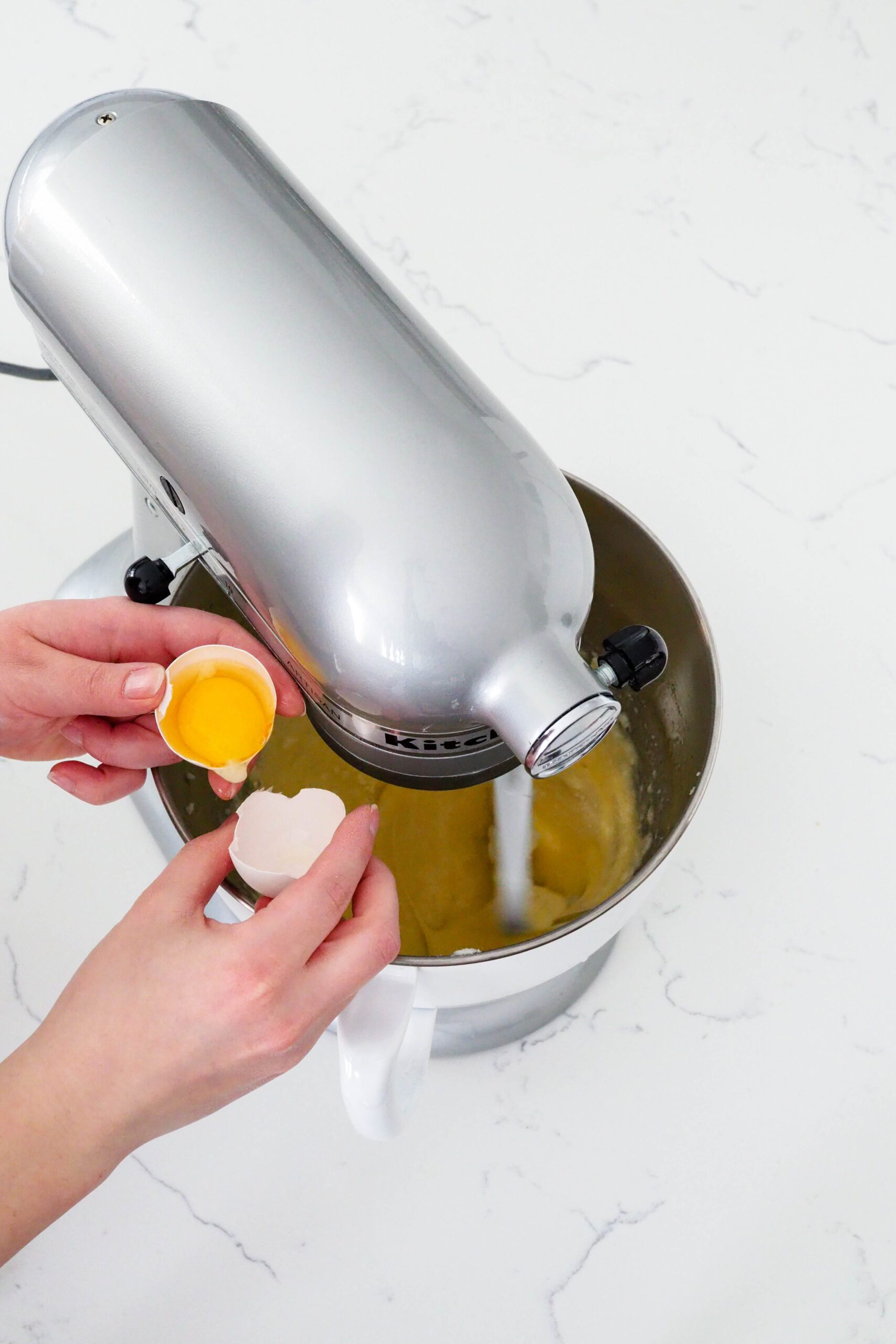 Two hands separate an egg into a stand mixer bowl.