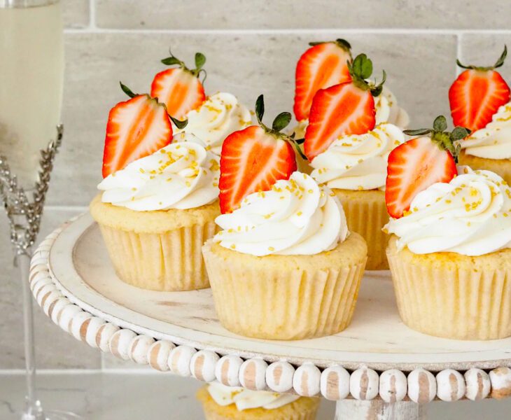 Strawberry champagne cupcakes on a distressed wooden cake stand with champagne glasses beside them.