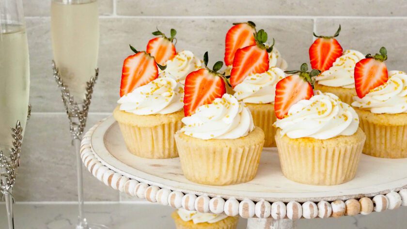 Strawberry champagne cupcakes on a distressed wooden cake stand with champagne glasses beside them.