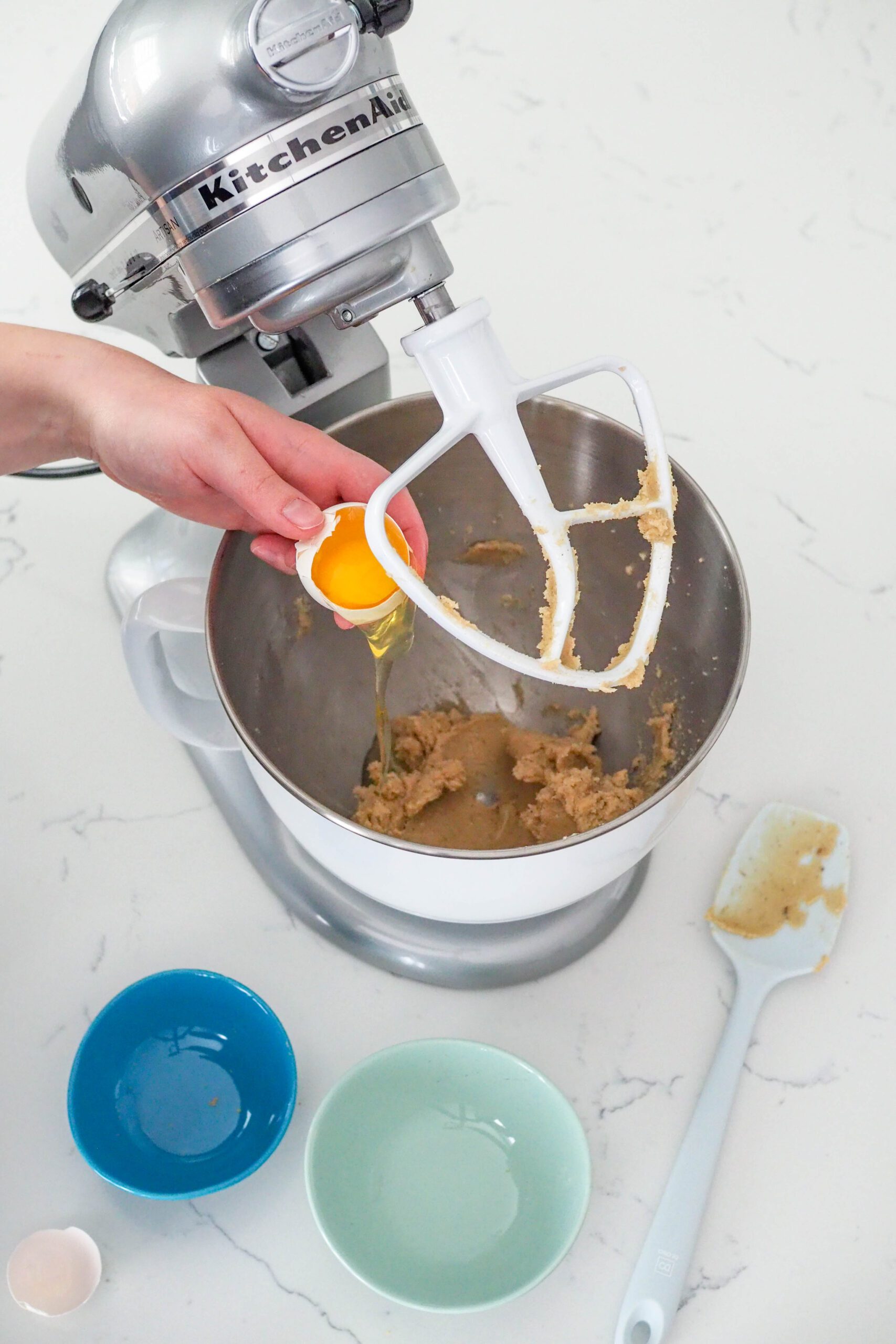 An egg that has been cracked open is held over the bowl of a stand mixer with cookie dough.