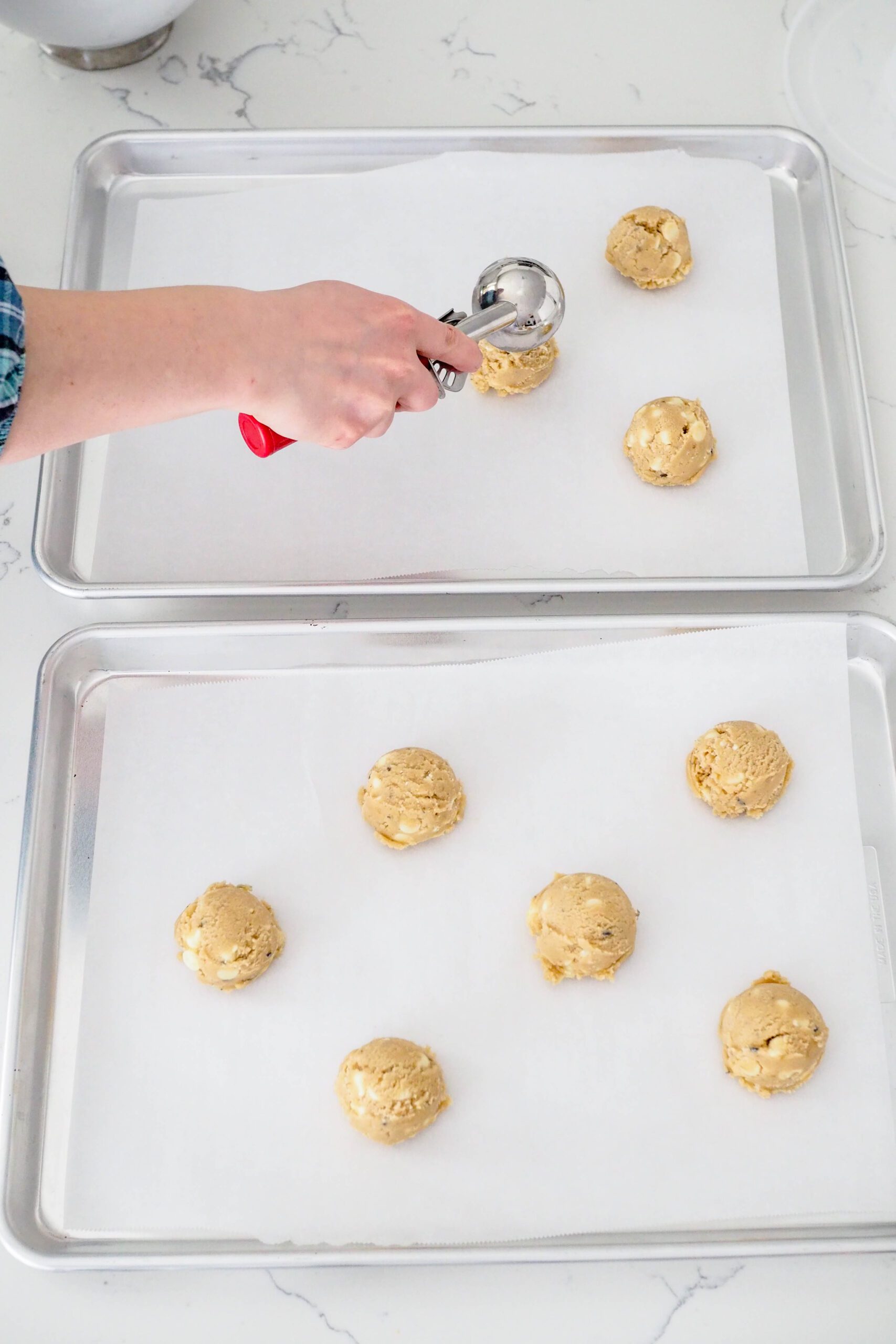 A hand scoops lavender chocolate chip cookie dough onto trays lined with parchment paper.