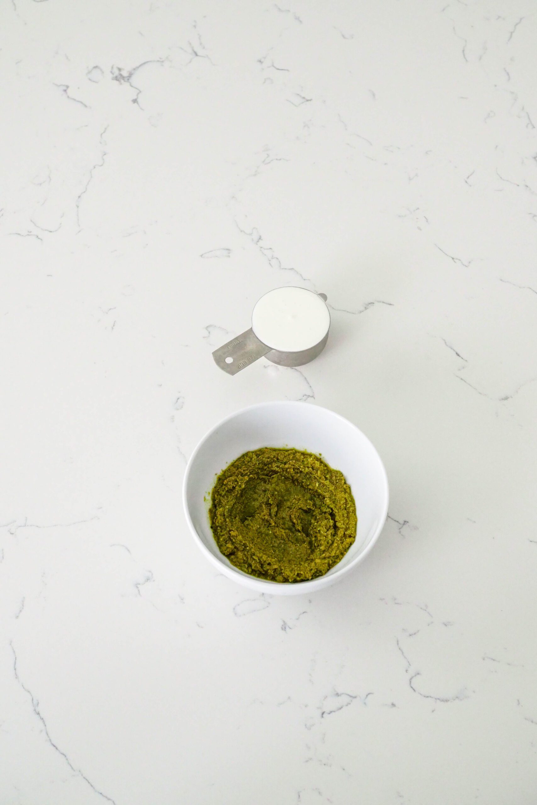 A bowl of pesto and a measuring cup full of heavy cream on a quartz counter.