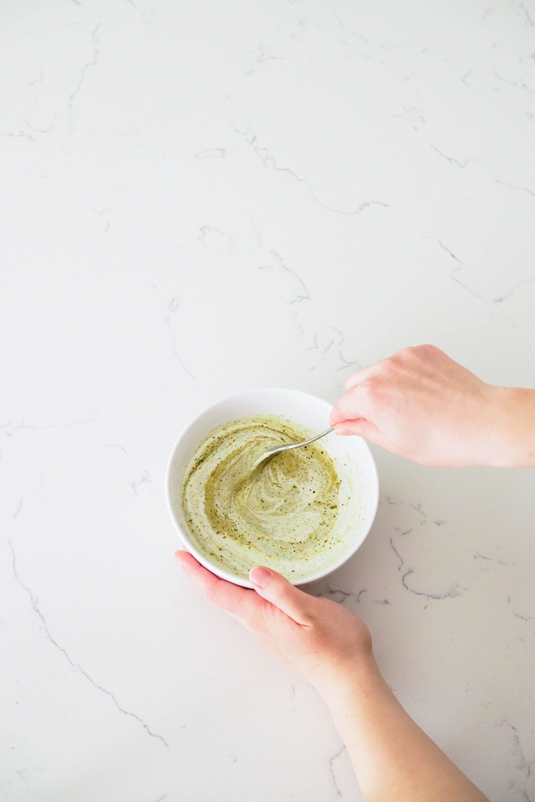 A hand stirs creamy basil pesto together in a bowl.