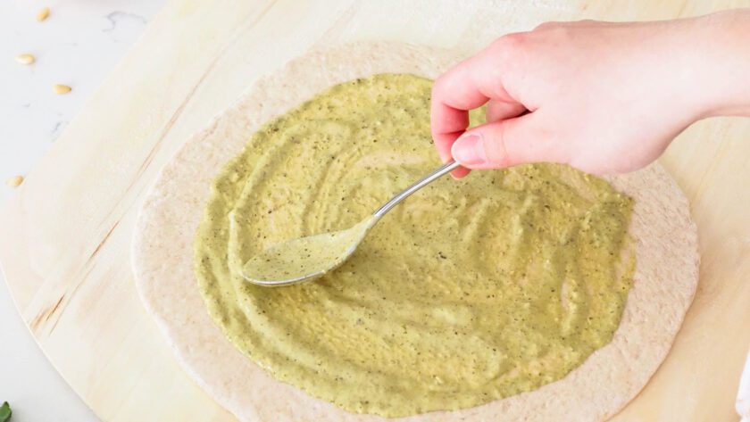 A hand spreads creamy basil pesto onto a round of pizza dough.
